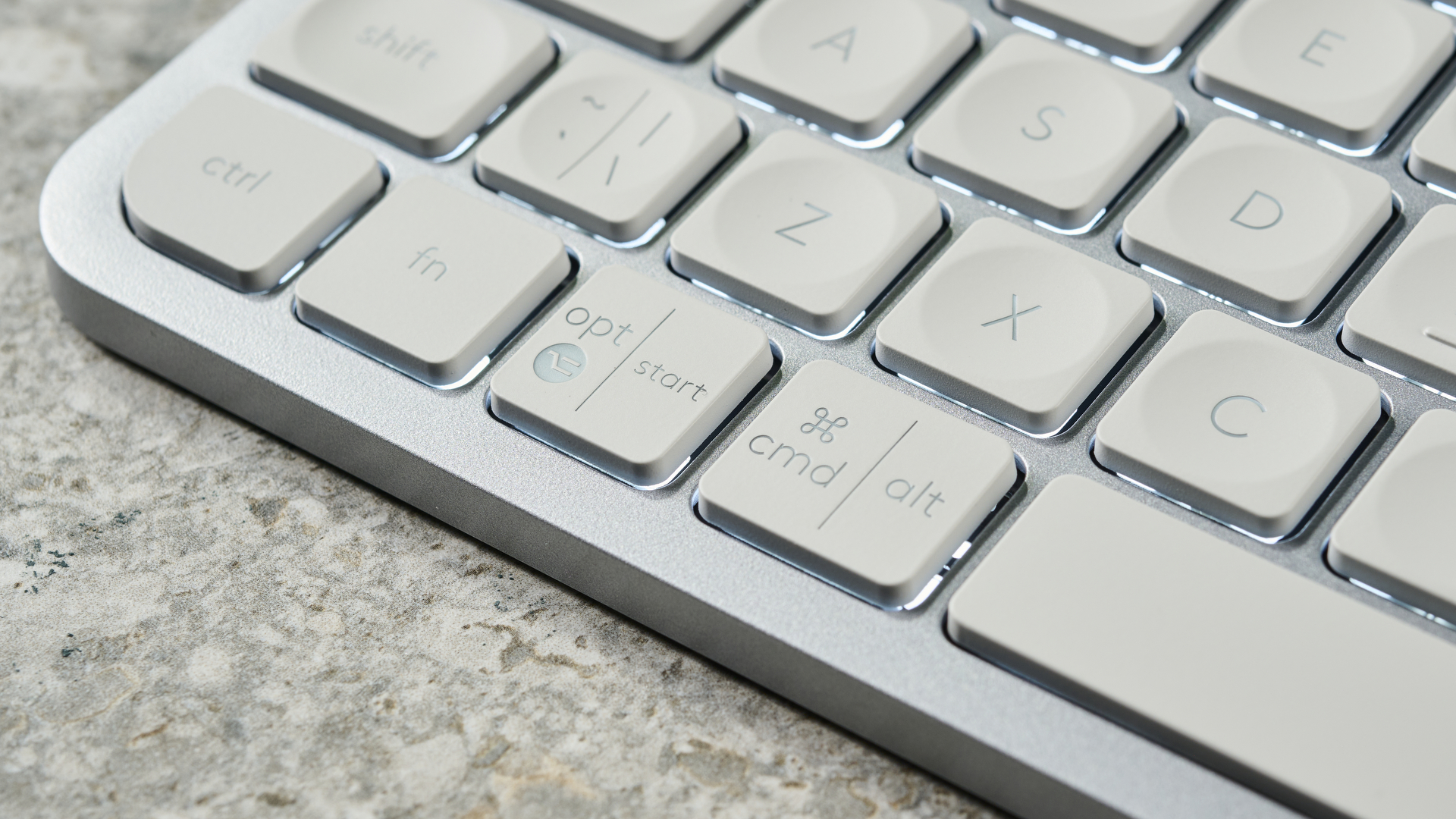 Una fotografia della Logitech MX Keys Mini in grigio chiaro, posizionata su una lastra di pietra con una parete blu sullo sfondo.